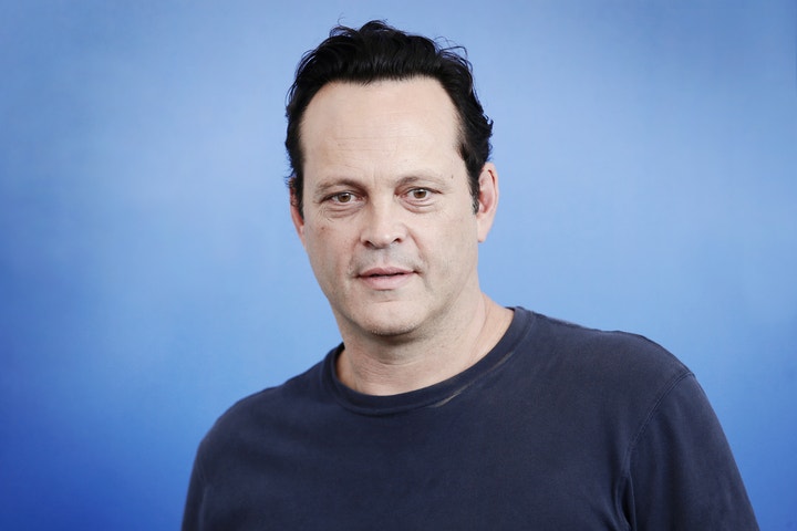 Actor Vince Vaughn stands in front of a financial chart, smiling.